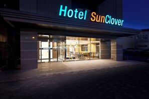 A hotel entrance with a sign reading 'Hotel SunClover,' glass doors, and a seating area outside.