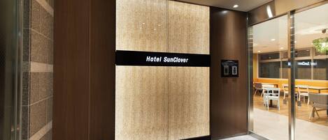 A hotel entrance with a modern design, featuring a large sign for Hotel SunClover, a glass door leading to an indoor area, and a tiled floor.
