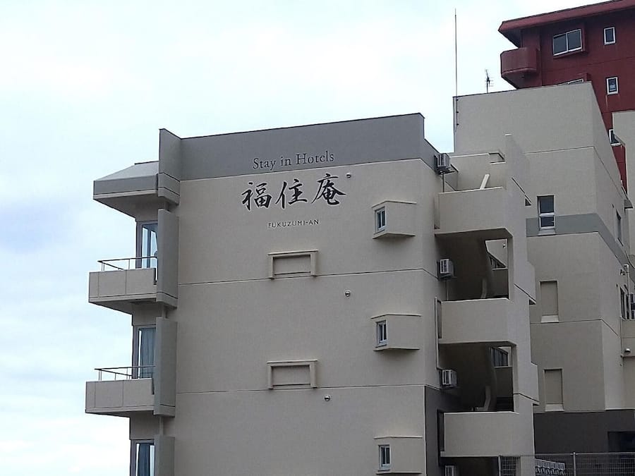 Ein mehrstöckiges Hotel mit Balkonen, einem Schild mit der Aufschrift 'Stay in Hotels' und dem Namen 'FUKUZUMI-AN' in englischer Schrift und einer weiteren Schrift.