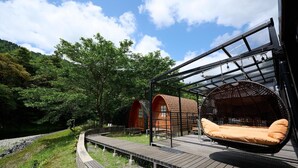 Terraza de madera con una silla colgante y un área con cerramientos de vidrio, rodeada de árboles y un cielo despejado.
