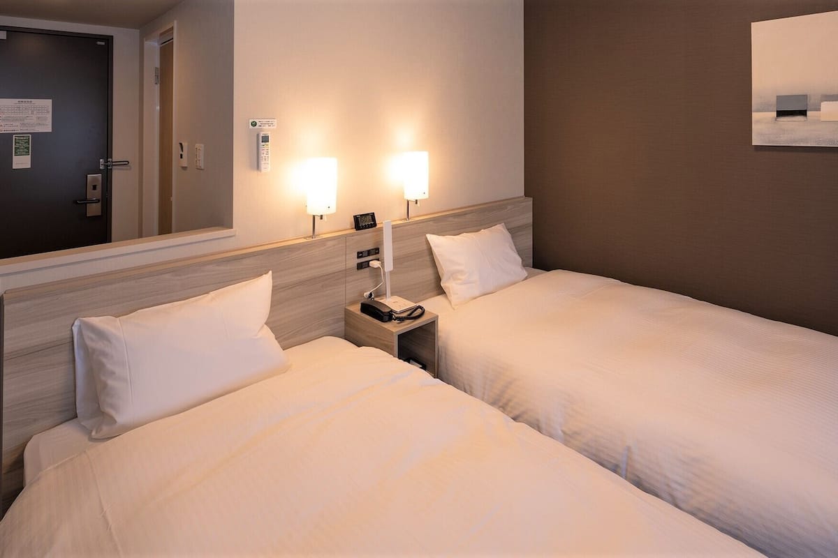 Habitación de hotel con dos camas, una mesita de noche con teléfono y lámpara, y un aplique de pared.