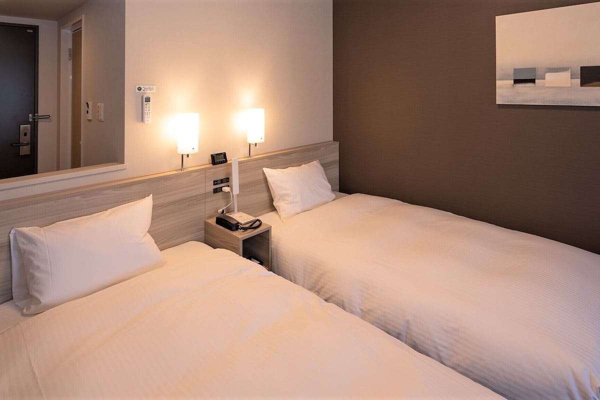 Habitación de hotel con dos camas, mesitas de noche con lámparas, un reloj de pared y un cuadro enmarcado.