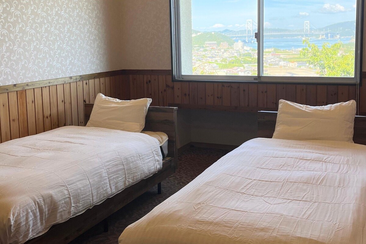 Una habitación con dos camas, un ventanal con vista a un puente y paisaje, paneles de madera y papel tapiz estampado.
