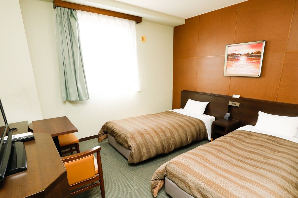 A hotel room with two beds, a desk with a computer, a chair, and a window with curtains.