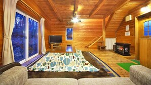A log cabin interior with a wooden floor, a fireplace, a television, and a large window with curtains.