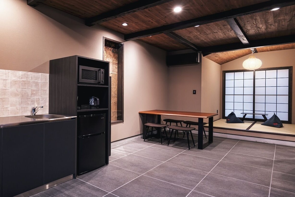 Uma cozinha com micro-ondas embutido, pia e geladeira preta. Há uma mesa de madeira com quatro cadeiras e uma janela com vista para o exterior.