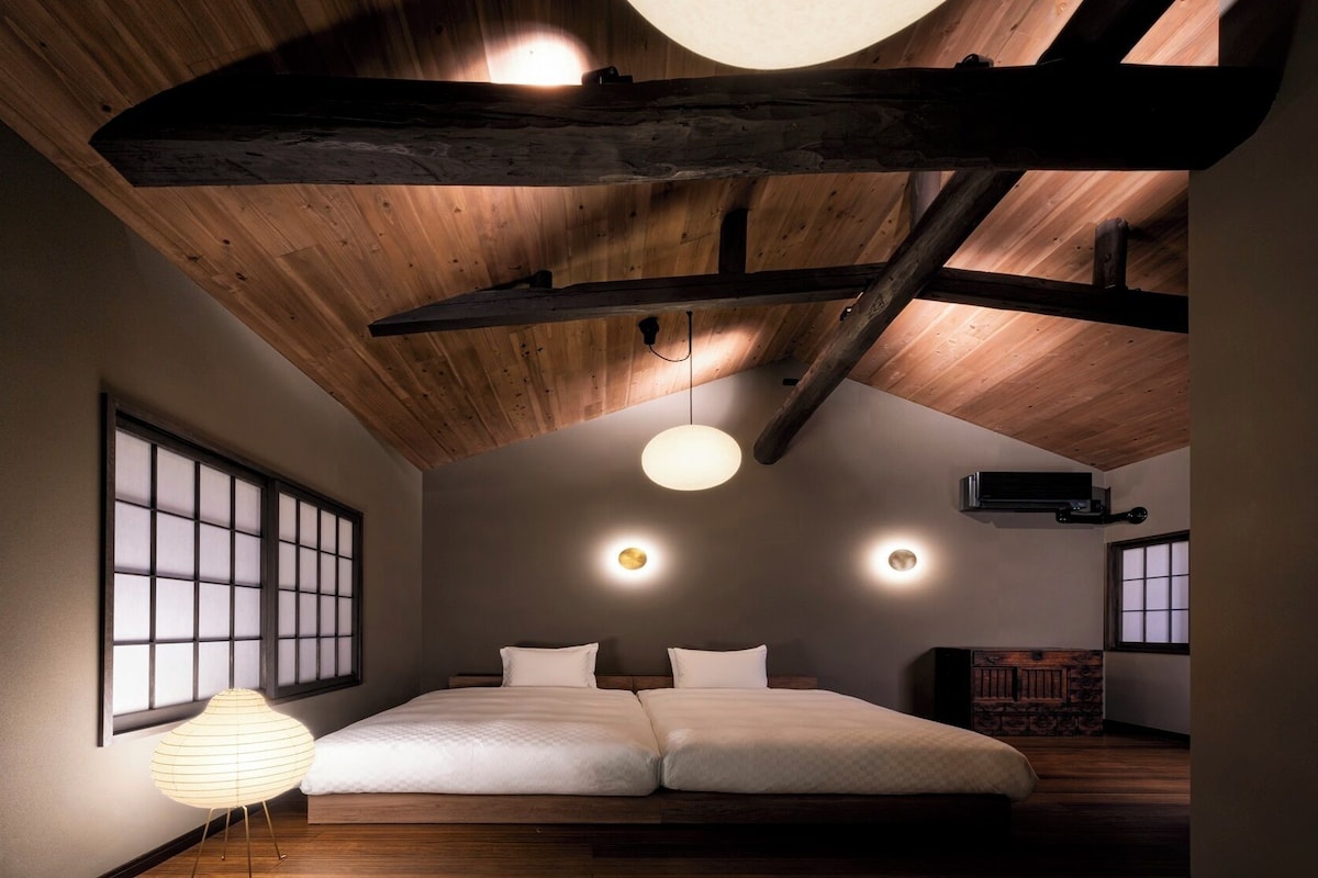 Quarto com teto inclinado, vigas de madeira, uma cama grande e um abajur de estilo tradicional japonês.