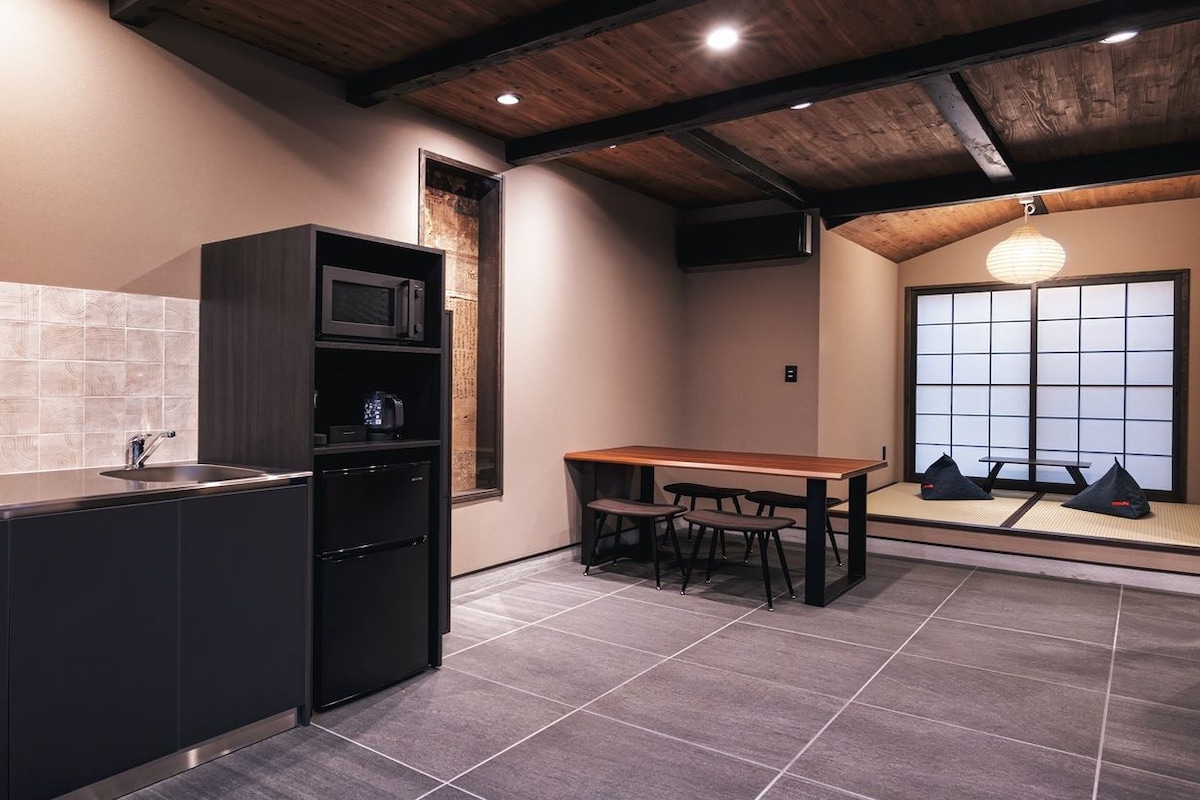 Uma cozinha com micro-ondas embutido, pia e geladeira preta. Há uma mesa de madeira com quatro cadeiras e uma janela com vista para o exterior.