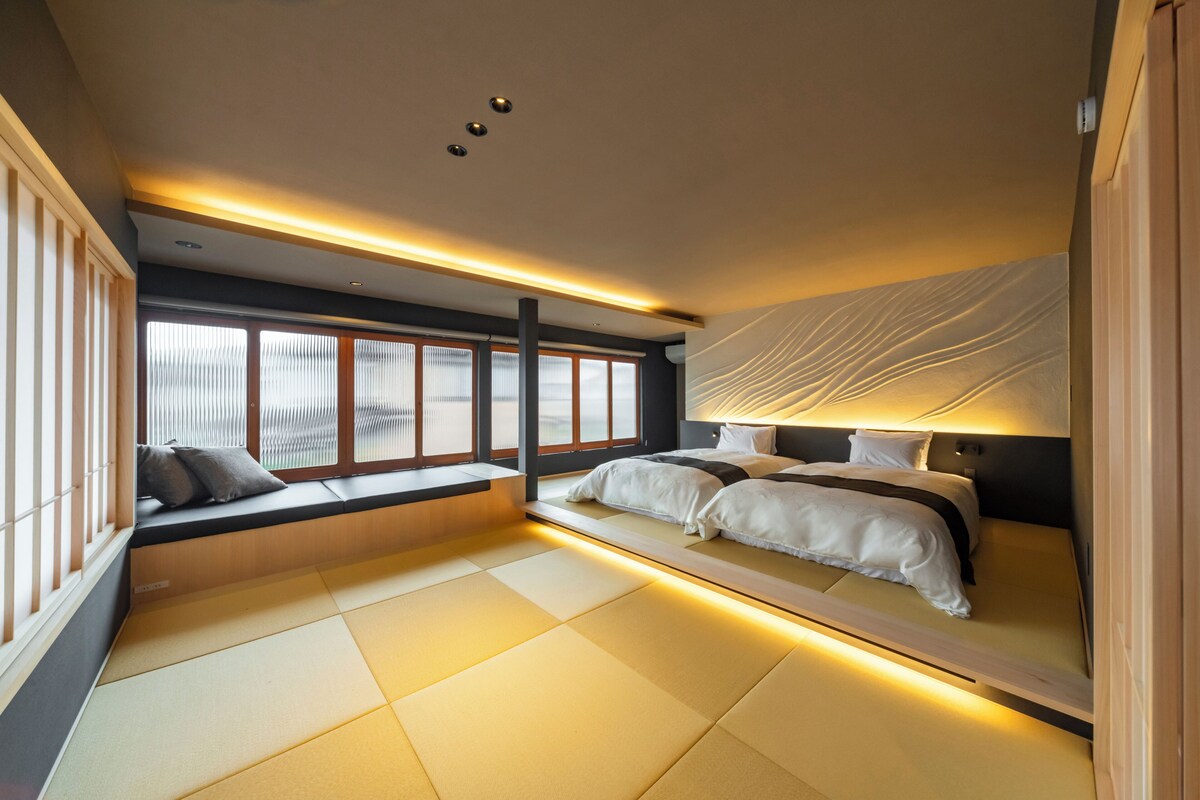 A modern hotel room with tatami flooring, a large bed, a window seat, and a wall-mounted headboard.