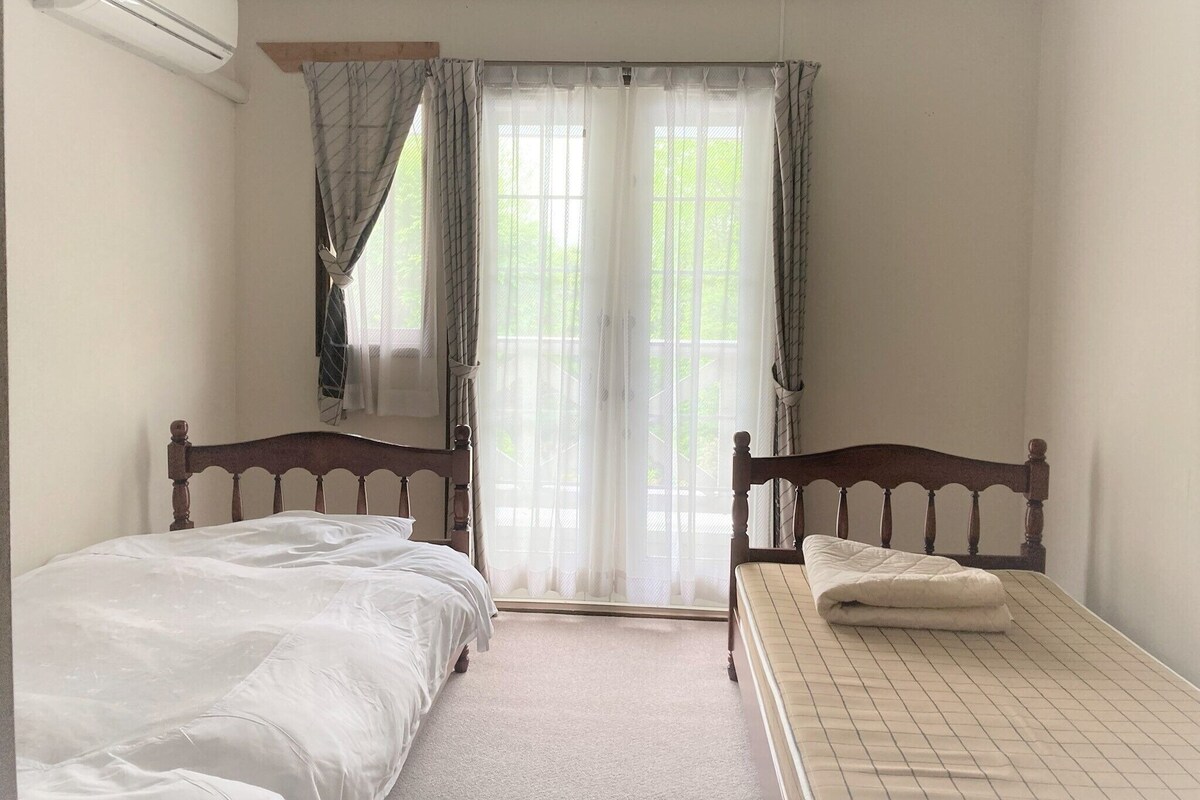 A bedroom with two wooden beds, a window with sheer curtains, and an air conditioning unit on the wall.