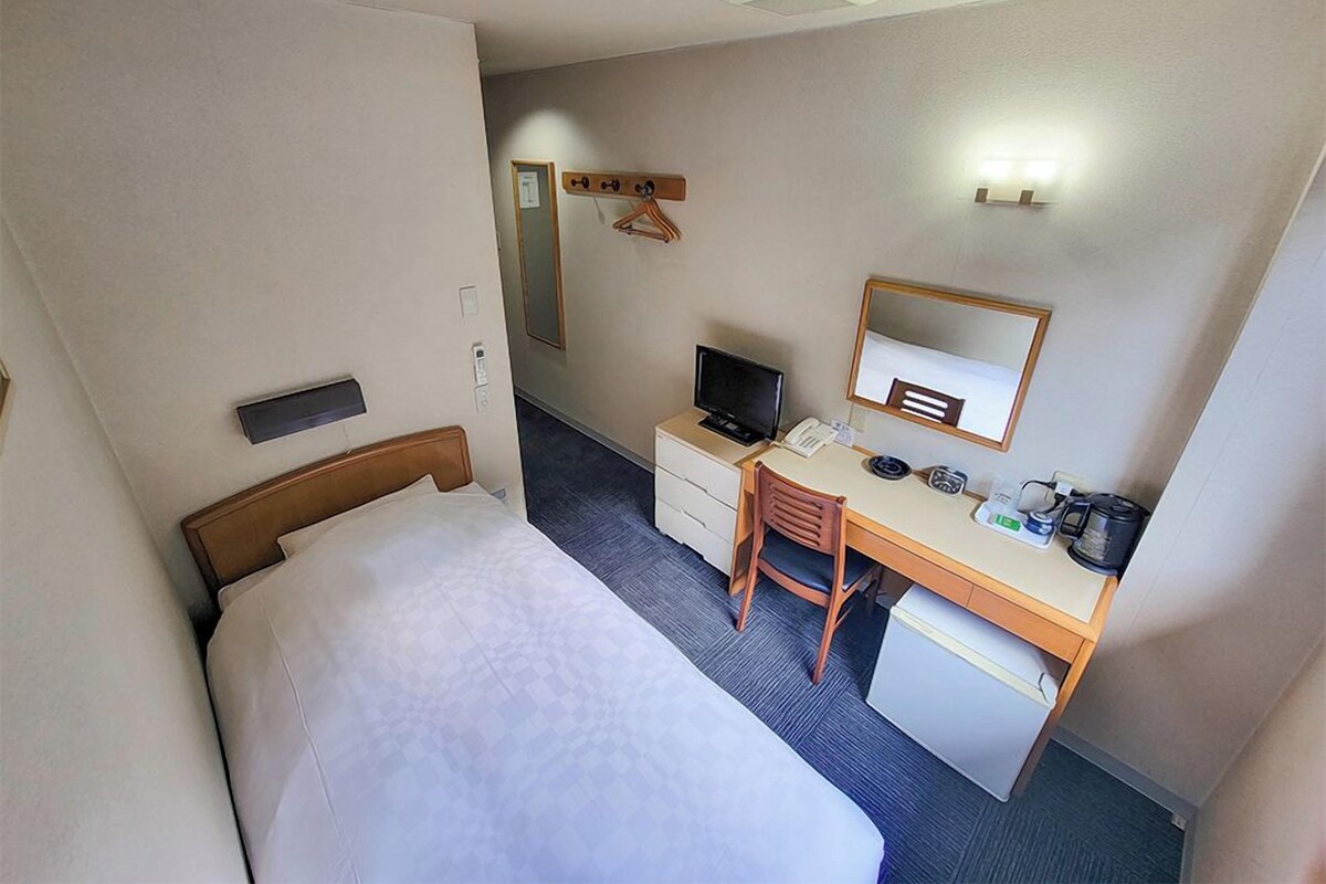 Habitación de hotel con cama, escritorio, silla, refrigerador pequeño, televisor y espejo.