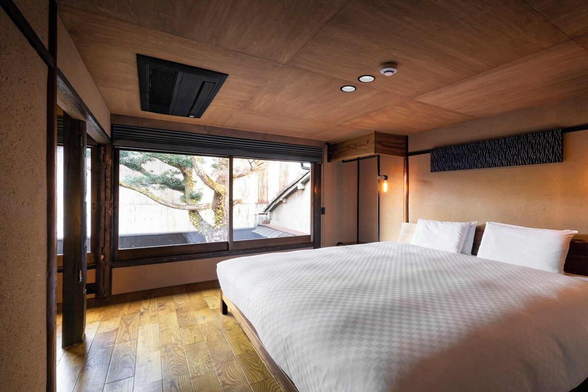 A bedroom with a large bed, wooden flooring, a window with a view, and a ceiling with recessed lighting.