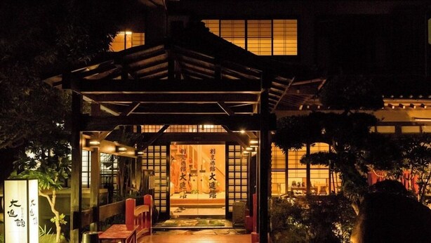A traditional Japanese building at night with a wooden structure in front, illuminated windows, and a sign with Japanese characters.