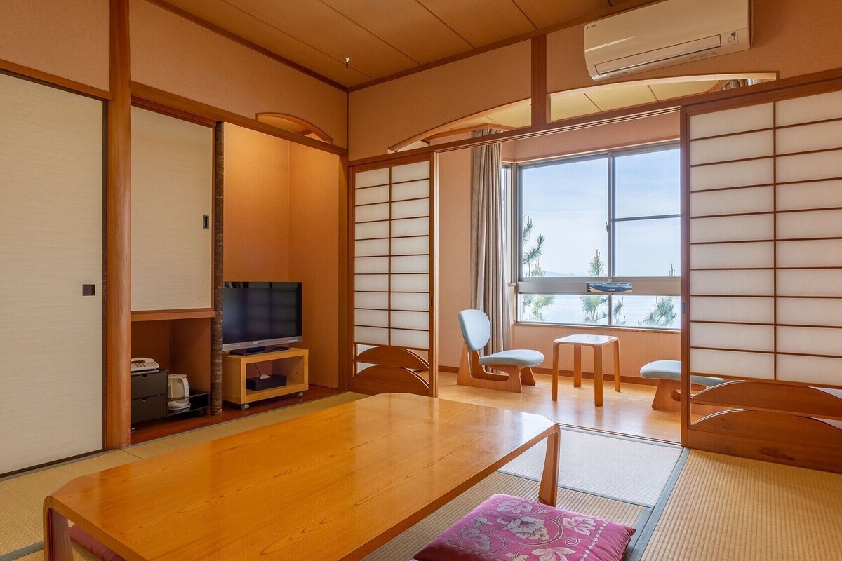 Une pièce de style japonais traditionnel, avec un sol recouvert de tatamis, une table basse, un téléviseur et une vue sur la mer, visible à travers des paravents en papier.