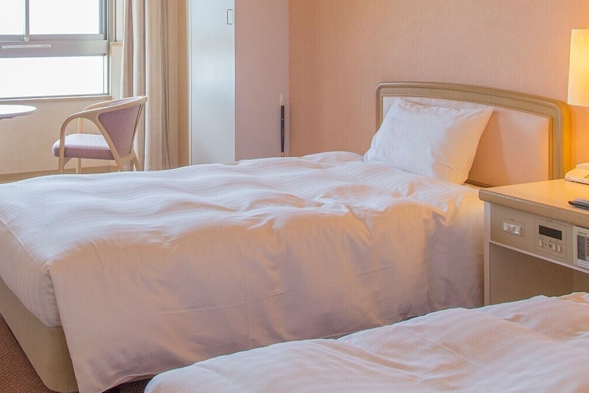 Une chambre d’hôtel avec un lit, une table de chevet, une lampe, une chaise et une fenêtre avec des rideaux.