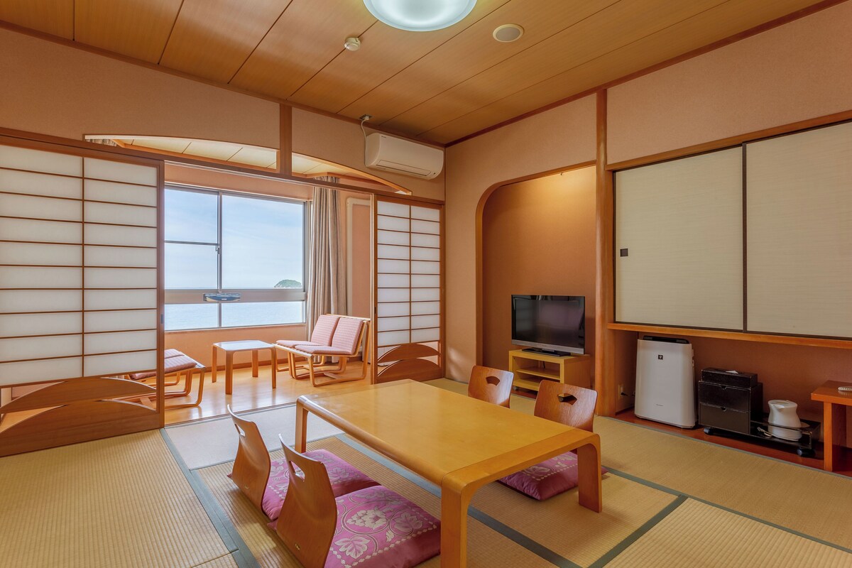 Une pièce de style japonais traditionnel, avec un sol recouvert de tatamis, une table basse, des chaises, un téléviseur et des portes coulissantes.