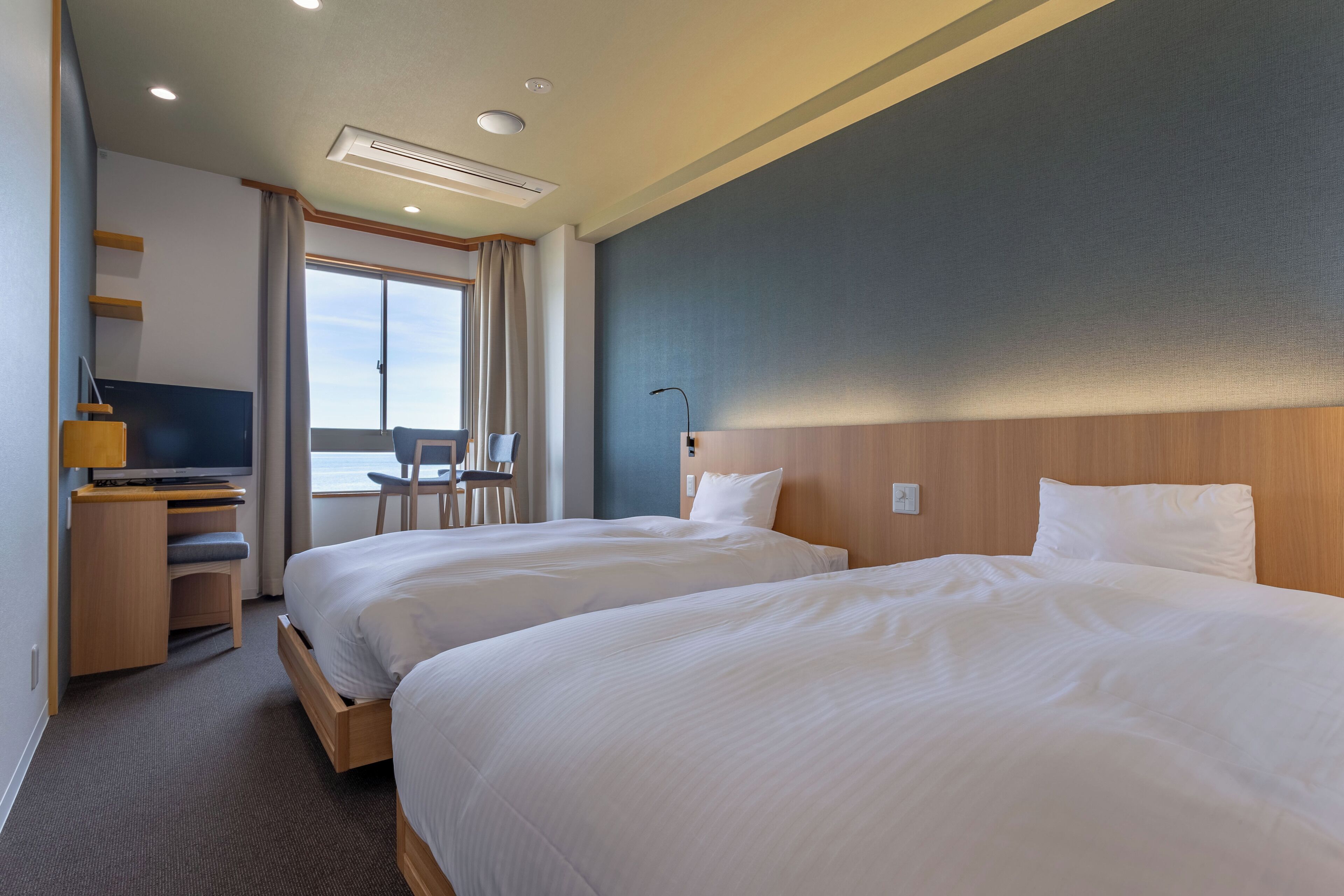 A hotel room with two beds, a desk, a chair, and a large window.