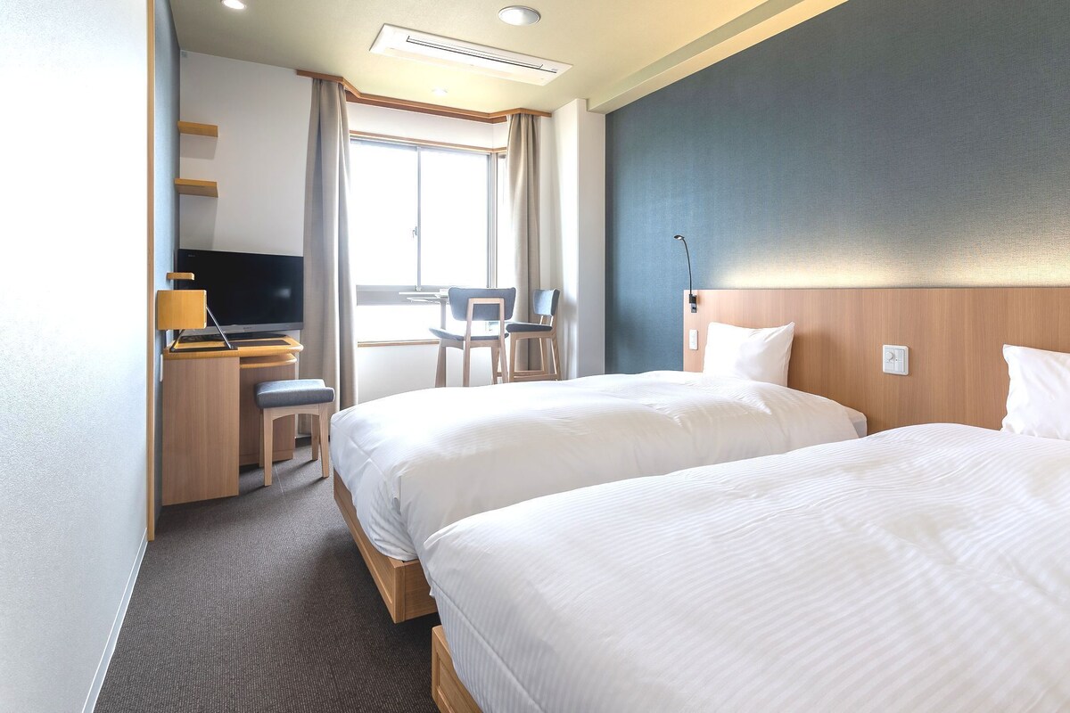 Une chambre d’hôtel avec deux lits, un bureau avec une chaise, une télévision et une grande fenêtre.