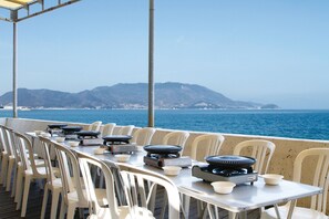 Ein Essbereich mit Tischen und Stühlen, Grillutensilien im Freien und Blick auf Meer und Berge.