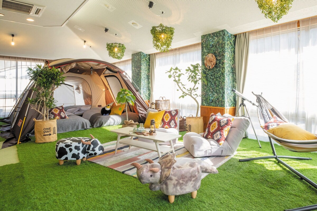 Un espace de camping intérieur accueillant, avec une tente, des sièges confortables, un hamac et un revêtement de sol en gazon synthétique.