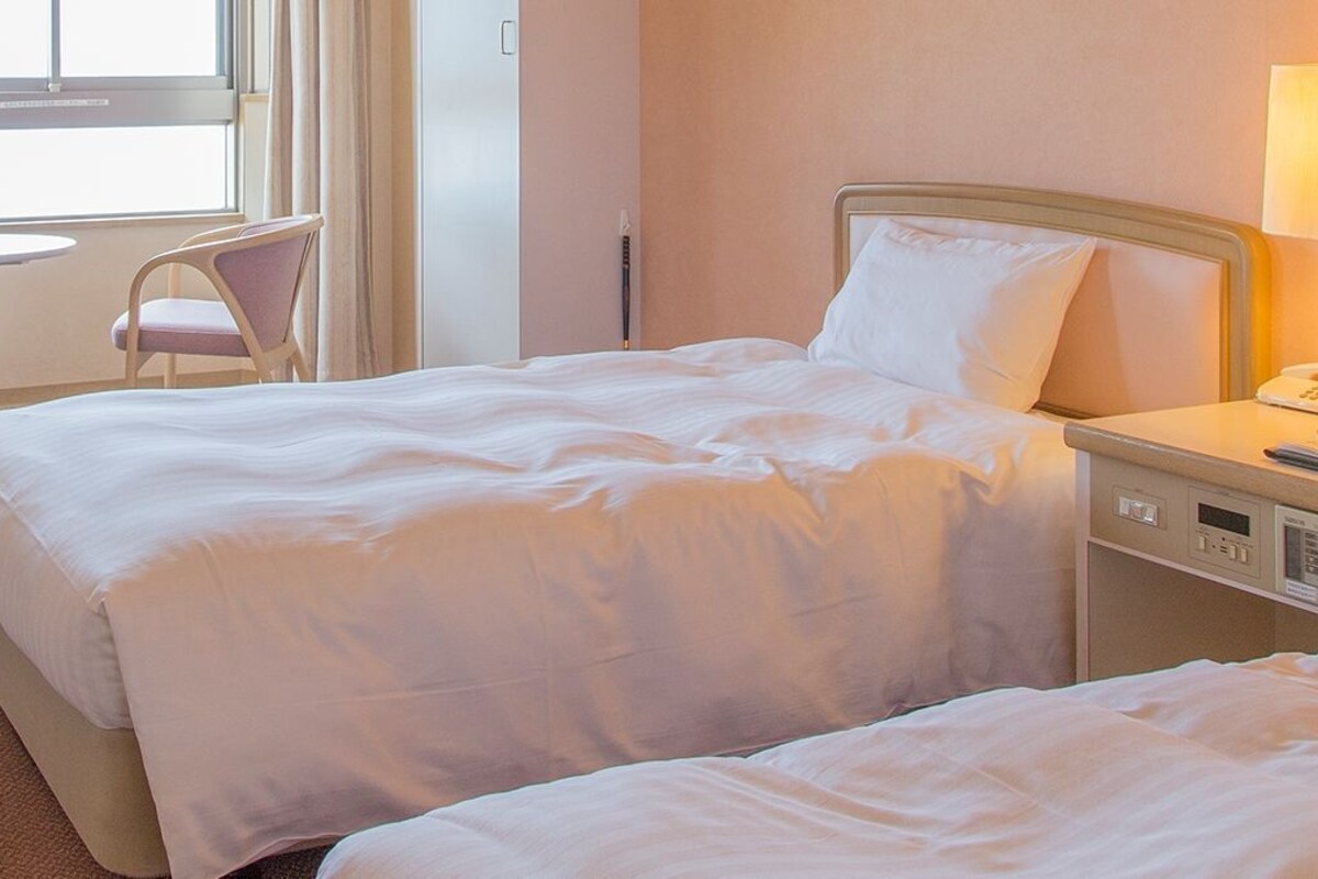 Une chambre d’hôtel avec un lit, une table de chevet, une lampe, une chaise et une fenêtre avec des rideaux.