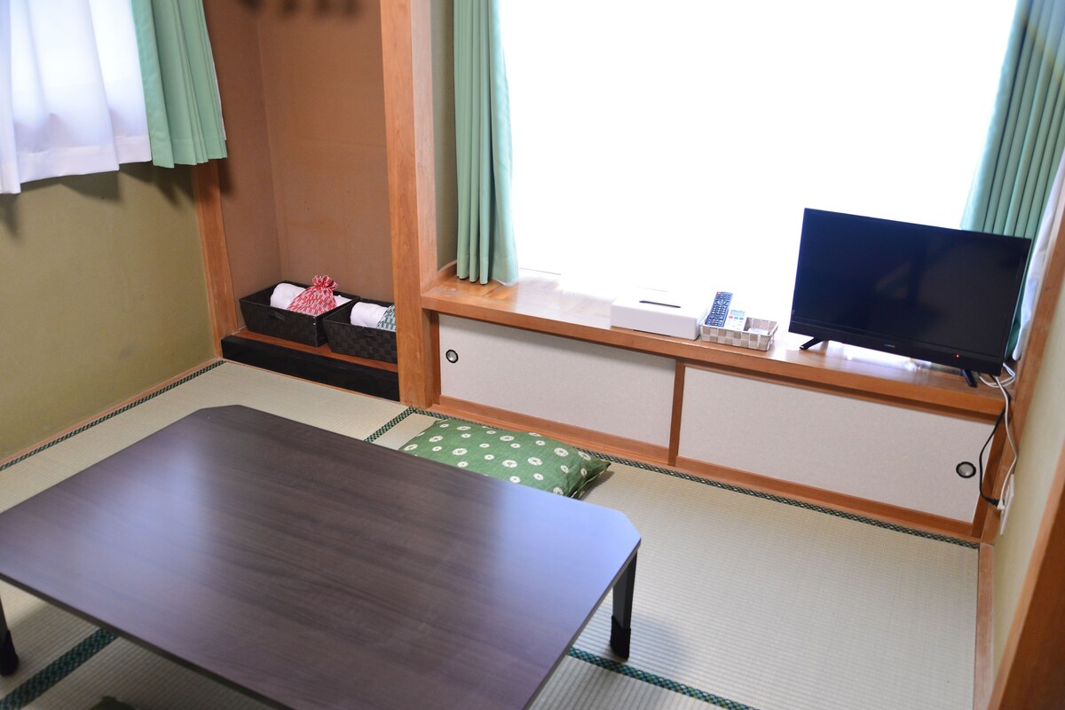 A room with a tatami mat floor, a low table, a television on a stand, a small shelf with items, and a window with curtains.