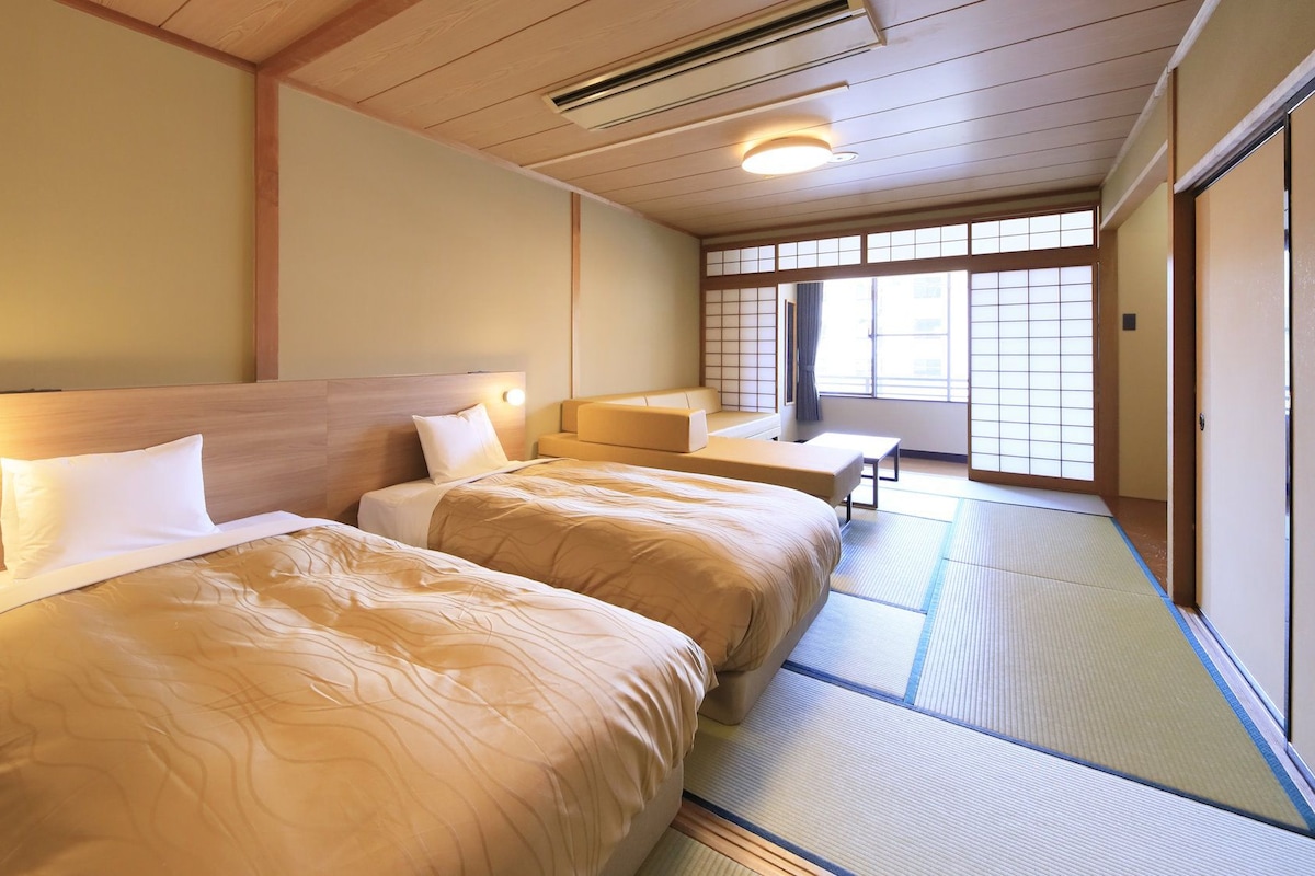 Habitación de hotel con dos camas, una mesita pequeña y vistas al exterior a través de amplios ventanales.