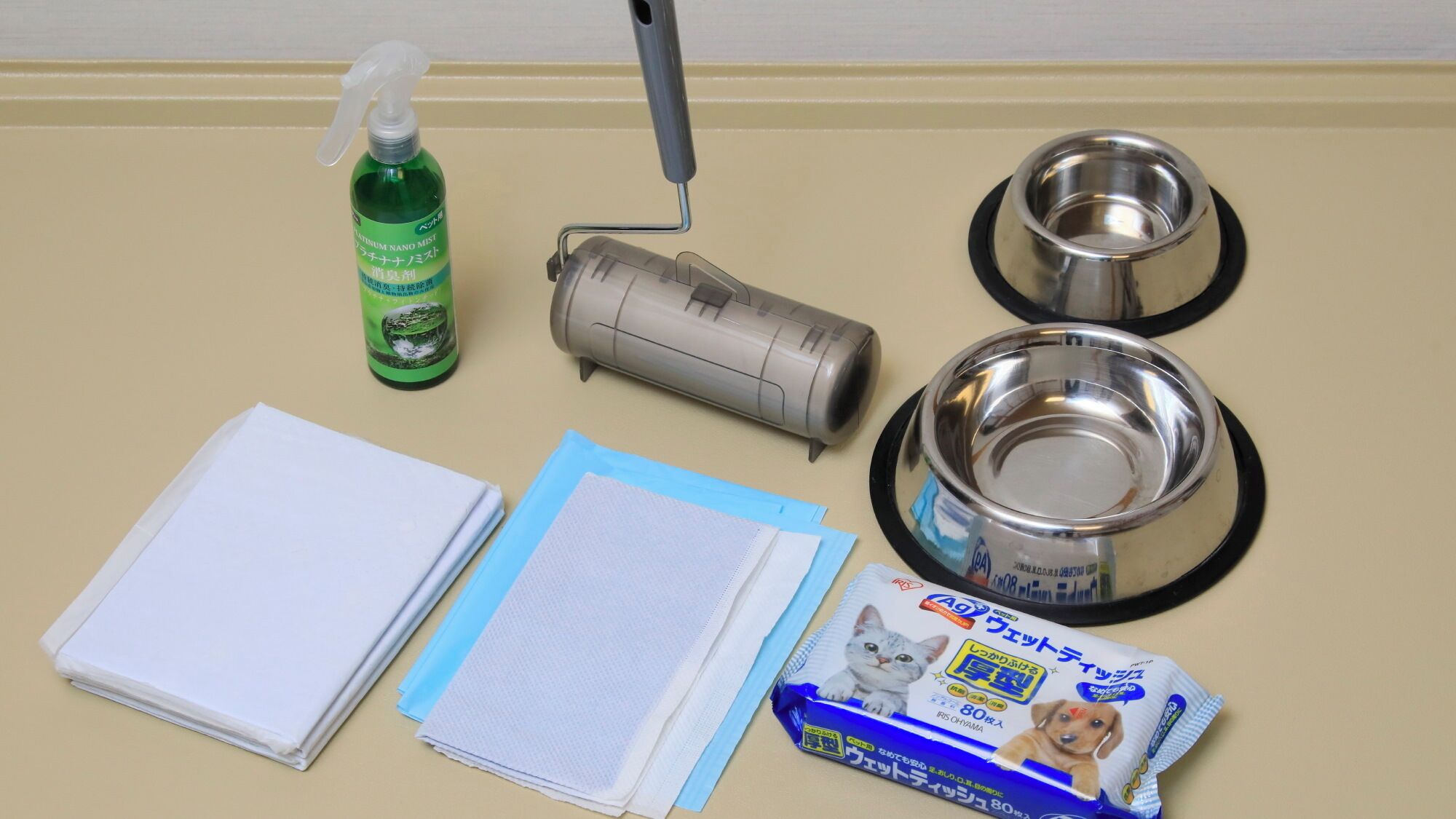 A table with a spray bottle, a paint roller, two metal bowls, a stack of white paper, a blue folder, and a package of cat litter.
