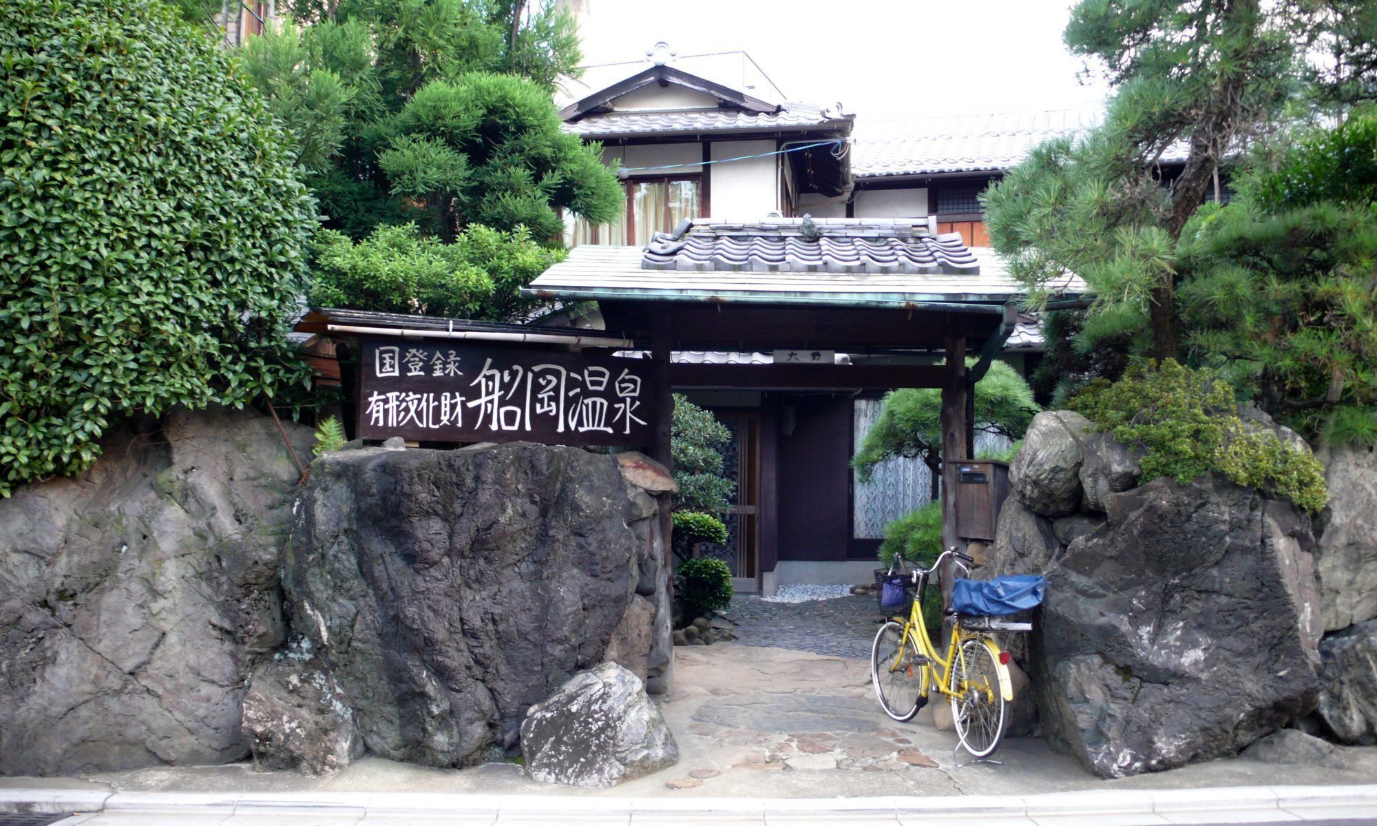 A traditional Japanese building with a tiled roof, a stone entrance gate, and a bicycle parked outside.