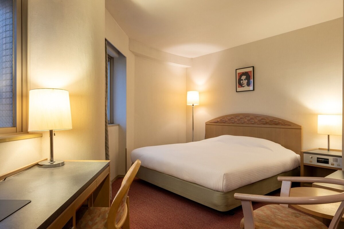 A hotel room with a bed, desk, chair, bedside lamps, and a picture on the wall.