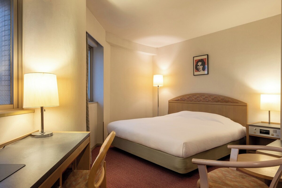 Una habitación de hotel con cama, escritorio, silla, lámparas de noche y un cuadro en la pared.