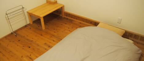 Un dormitorio con piso de madera, una cama individual, una mesita de noche de madera, una pequeña mesa y una ventana.