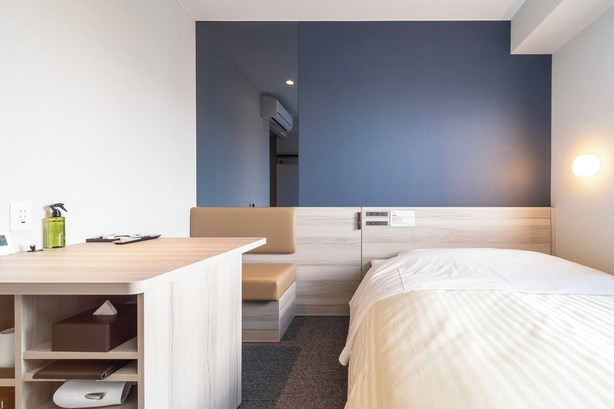 A hotel room with a bed, a desk, a chair, a small table, a shelf, a mirror, a wall-mounted air conditioner, a window, and a wall-mounted light.