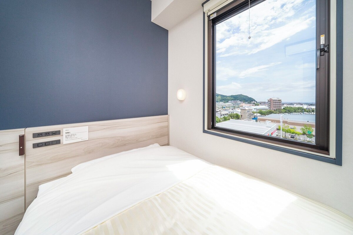 Habitación de hotel con cama, ventana que ofrece vistas a un paisaje urbano y un enchufe en la pared.
