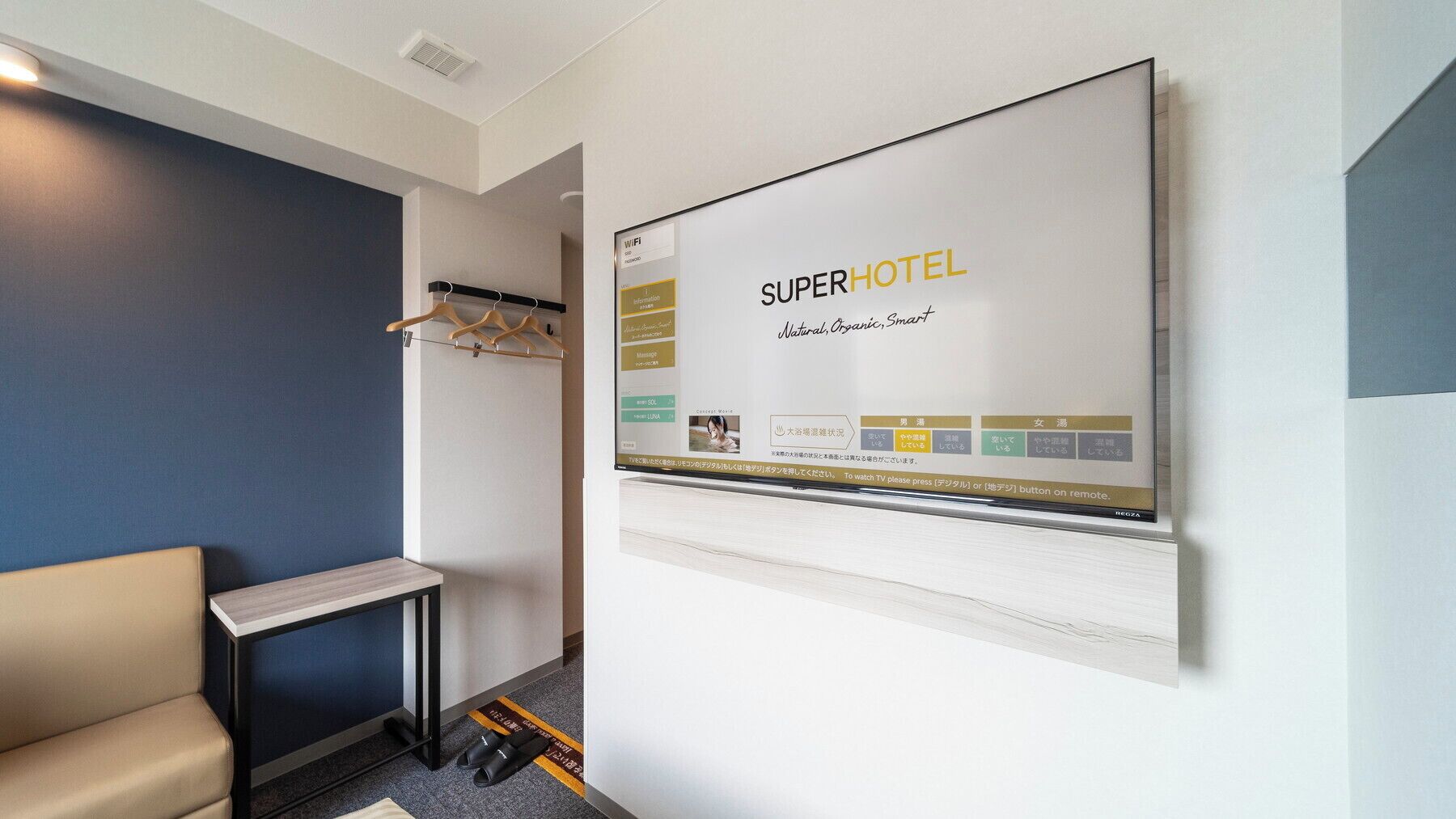 Une chambre d’hôtel dotée d’un grand écran numérique affichant les marques « SUPERHOTEL » et « Naturel, biologique, intelligent ». On y trouve une petite table avec un ordinateur portable, un porte-manteau avec des cintres et une paire de chaussures posées sur le sol.