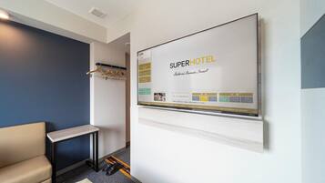 Une chambre d’hôtel dotée d’un grand écran numérique affichant les marques « SUPERHOTEL » et « Naturel, biologique, intelligent ». On y trouve une petite table avec un ordinateur portable, un porte-manteau avec des cintres et une paire de chaussures posées sur le sol.