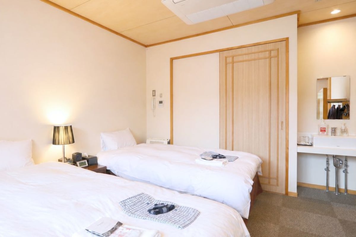Een hotelkamer met twee bedden, een nachtkastje, een lamp, een kledingkast en een badkamer met een wastafel en spiegel.