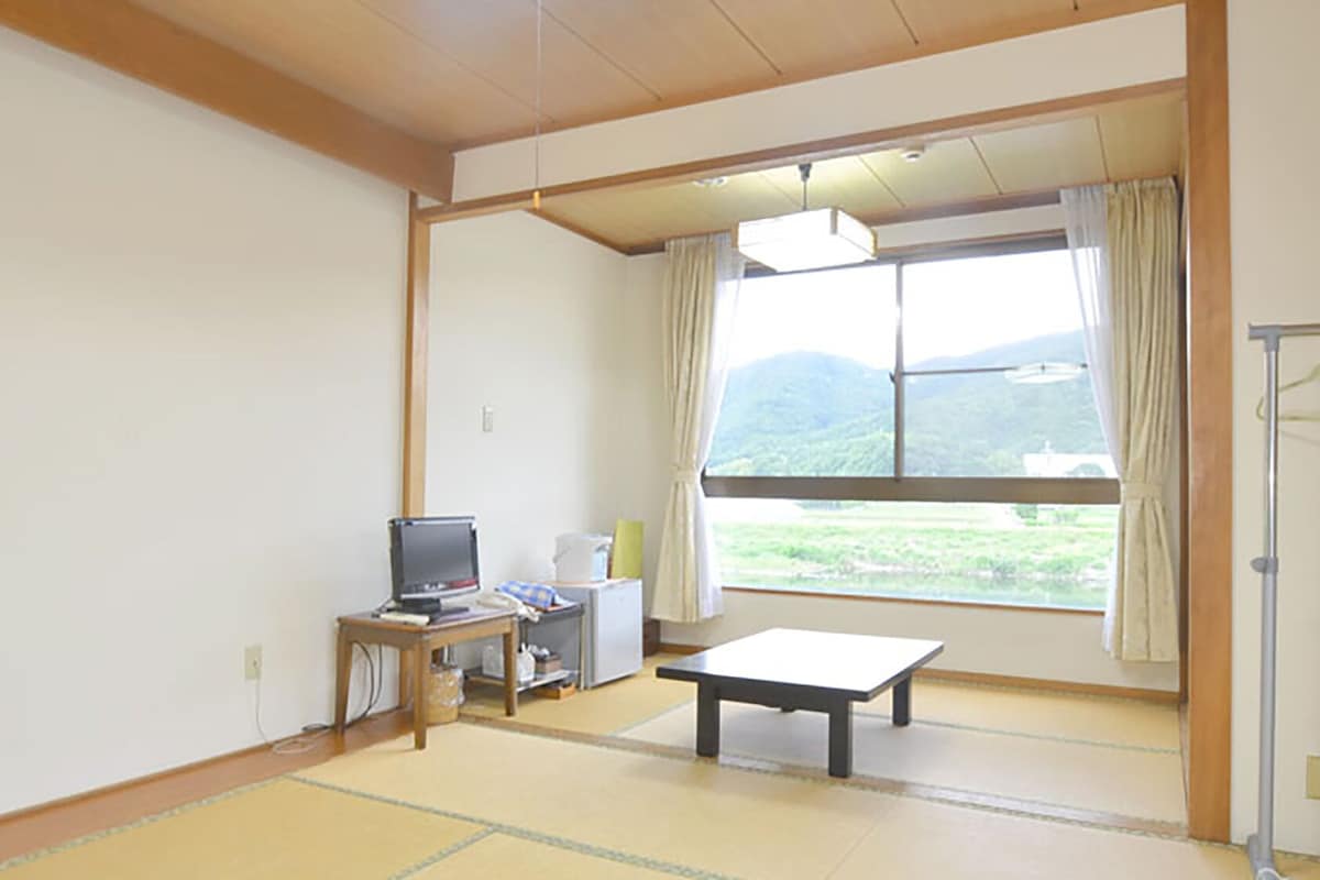 A room with tatami flooring, a desk with a computer, a table, and a view of a green landscape through large windows.