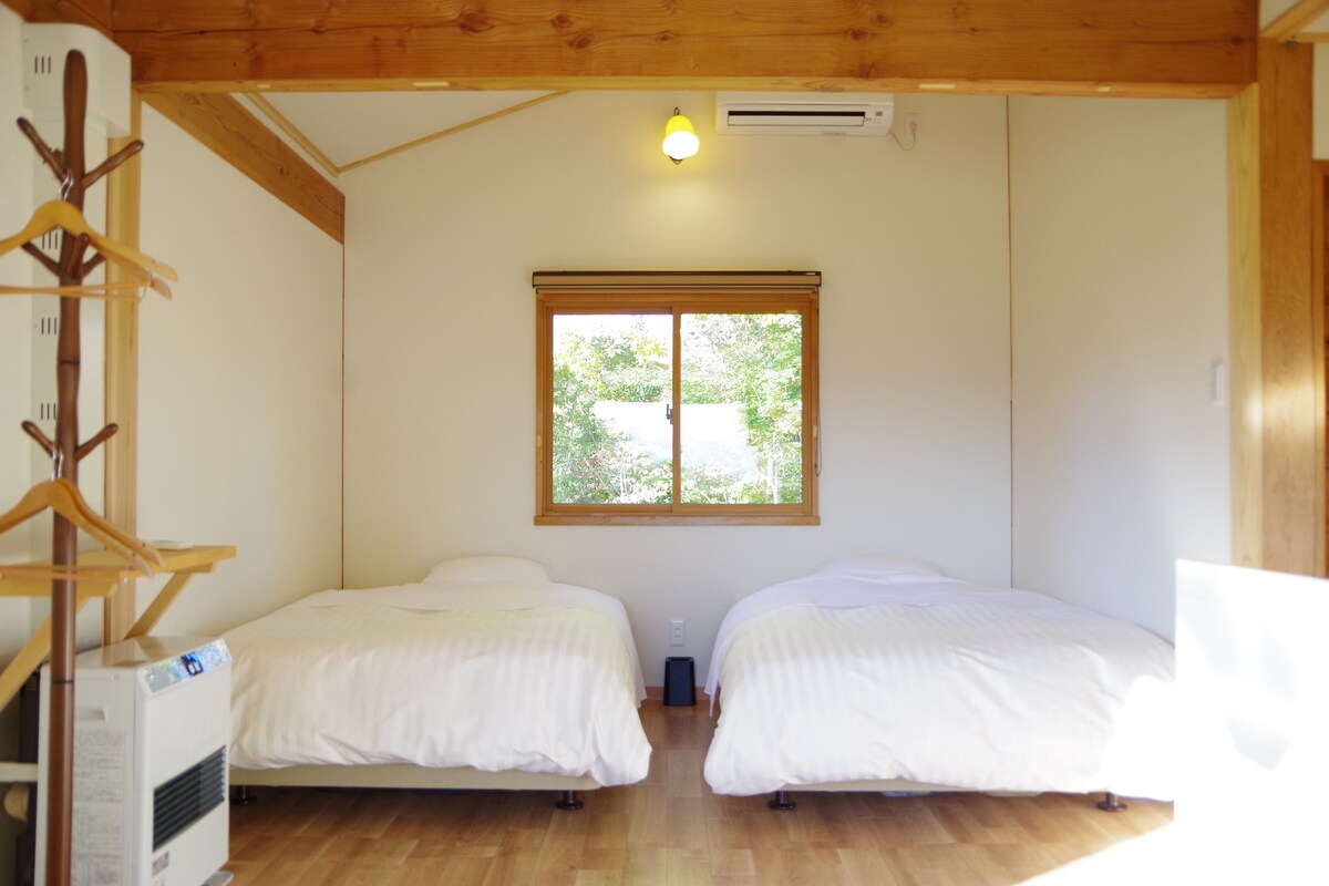 Una habitación con dos camas, suelo de madera, una ventana con vistas, un equipo de aire acondicionado y un perchero.