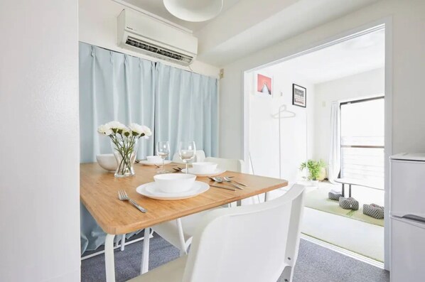 A dining area with a wooden table set for two, white chairs, a vase with white flowers, and a white refrigerator in the background. - Tora Hotel Kitaueno (Tokyo)
