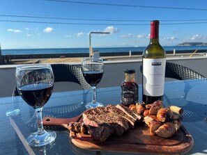 Uma mesa com um copo de vinho tinto, uma garrafa de vinho, um frasco de molho, e carne grelhada com legumes.