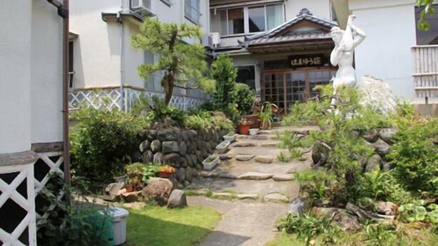 Ein traditionelles japanisches Haus mit einer Steintreppe, die zu einer Holztür führt, umgeben von einem Garten mit Steinen und Pflanzen.