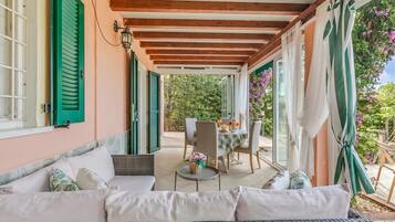 Un portico coperto con un divano in vimini, un tavolo da pranzo con sedie e vista sul giardino.