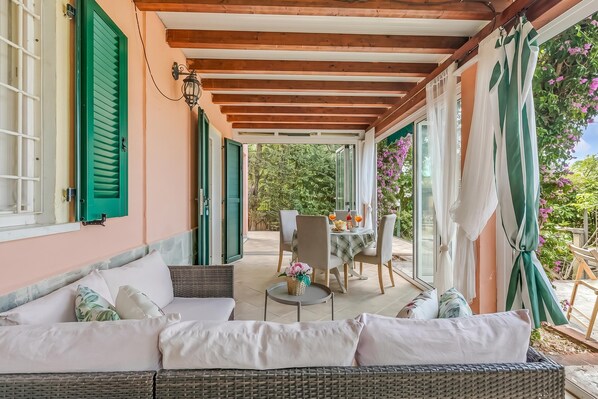 Un portico coperto con un divano in vimini, un tavolo da pranzo con sedie e vista sul giardino.