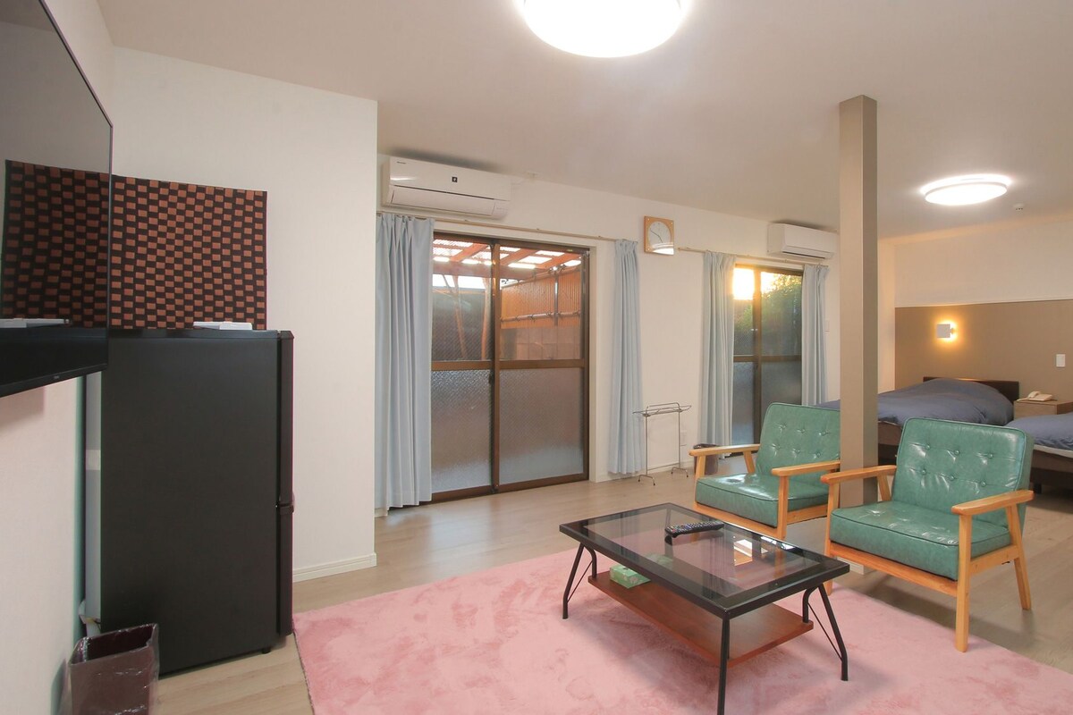 A hotel room with a bed, two chairs, a small table, a TV, and an air conditioning unit.