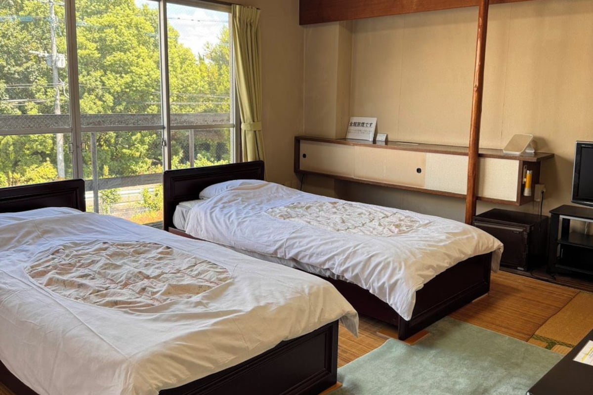 A hotel room with two beds, a desk, a television, and a window with a view of trees.