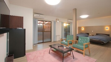 A hotel room with a bed, two chairs, a small table, a TV, and an air conditioning unit.