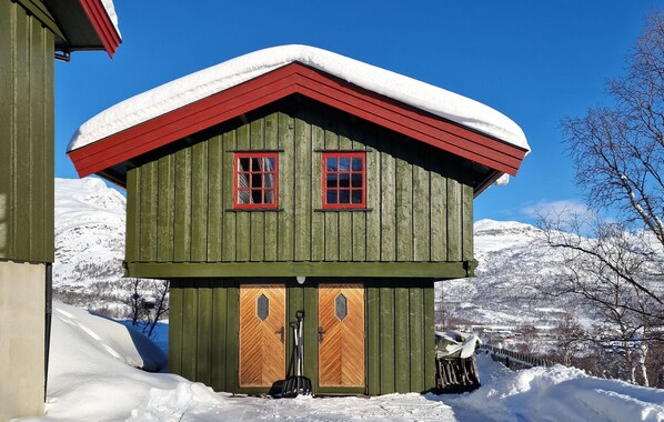 Una cabaña de madera con dos puertas y dos ventanas, rodeada de montañas nevadas.