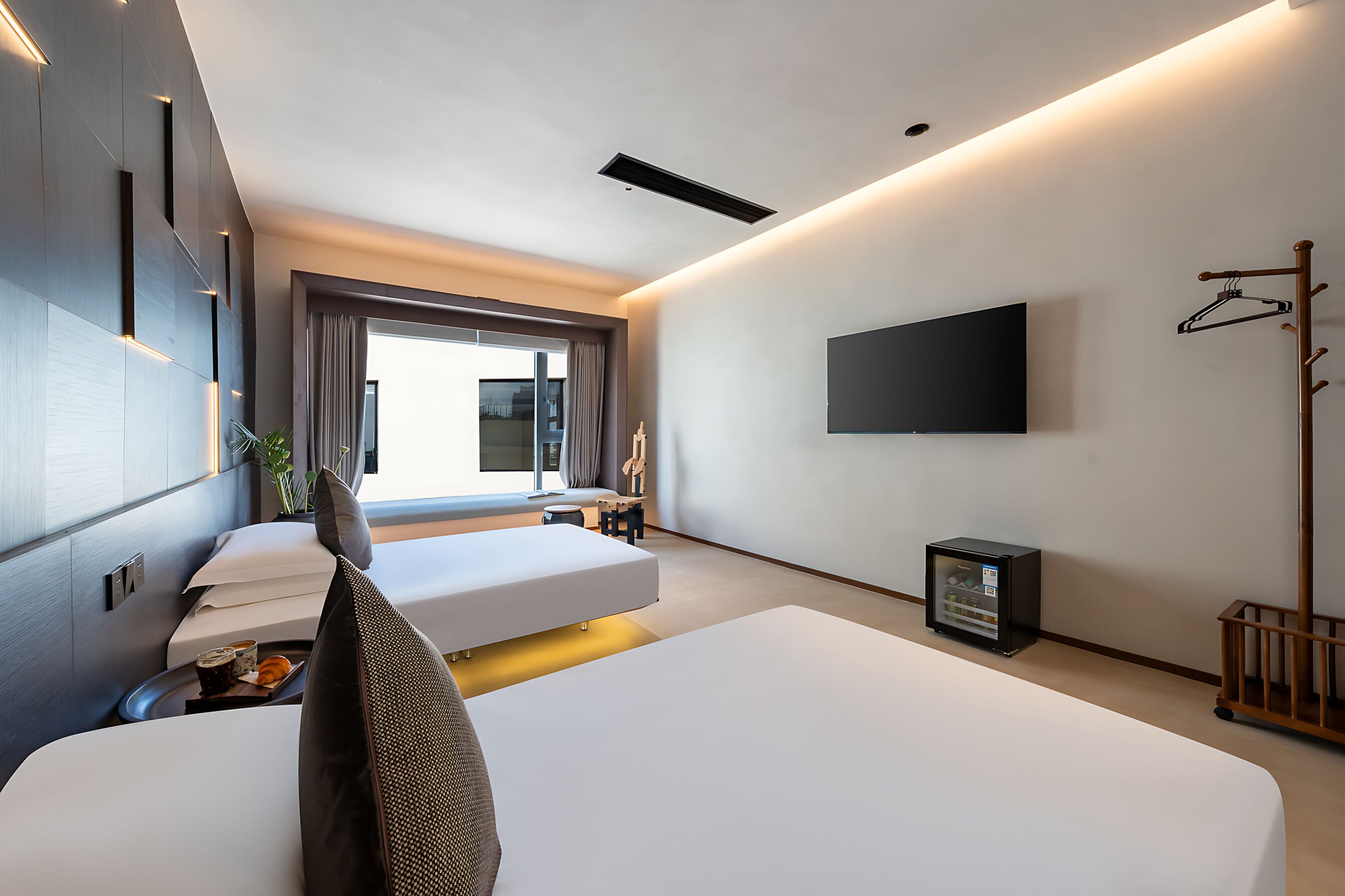 A modern hotel room with two beds, a flat-screen TV, a coat rack, and a small table with a plant.