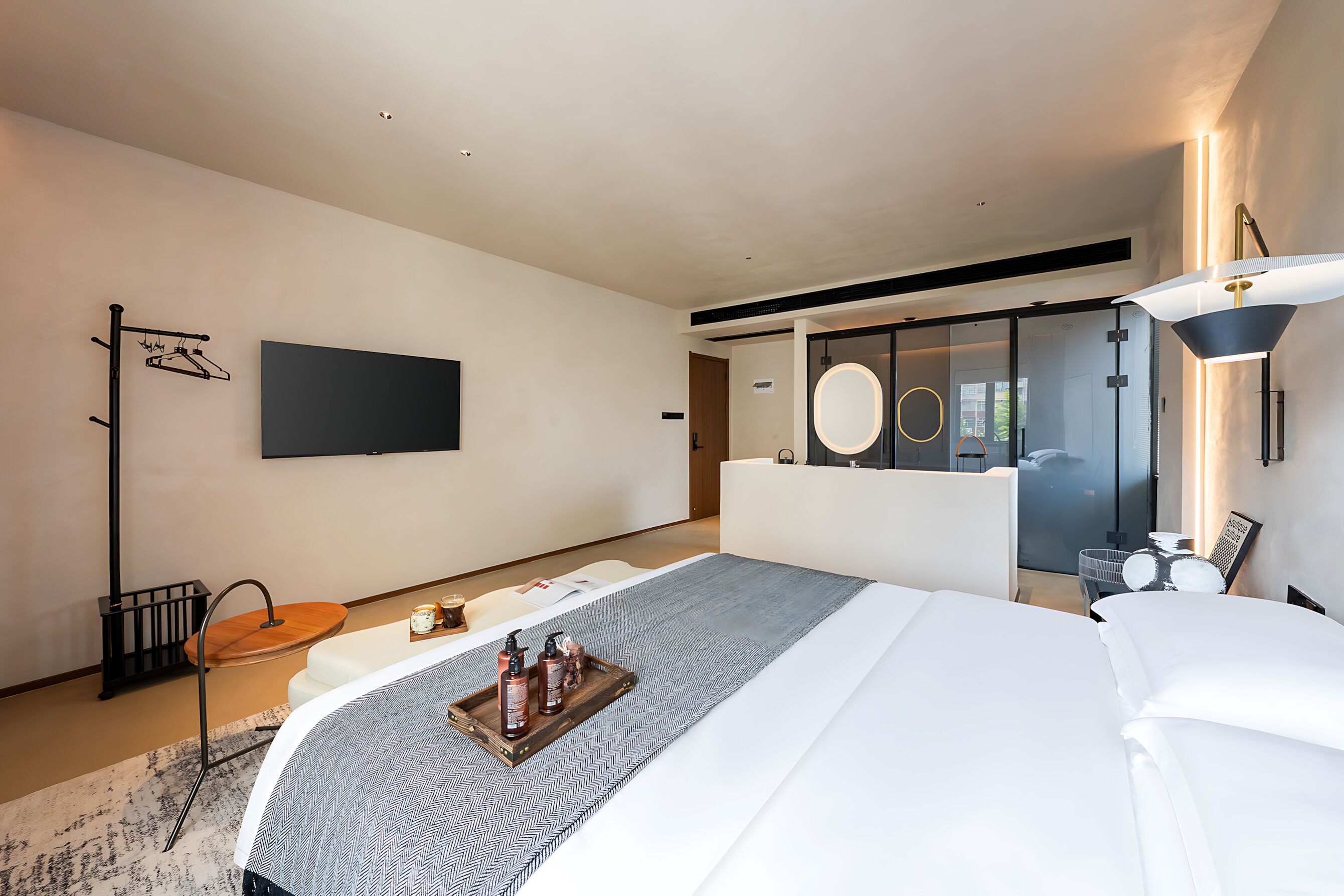 Ein modernes Hotelzimmer mit einem großen Bett, einem Nachttisch mit Tabletts für Toilettenartikel, einem an der Wand montierten Fernseher und einer gläsernen Duschkabine.